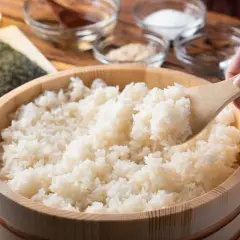 Drewniana miska z ryżem do sushi, nabierana drewnianą łyżką. W tle składniki.
