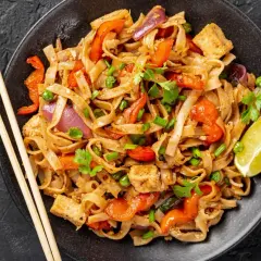 Pad thai z tofu, warzywami i limonką na czarnym talerzu, obok pałeczki.
