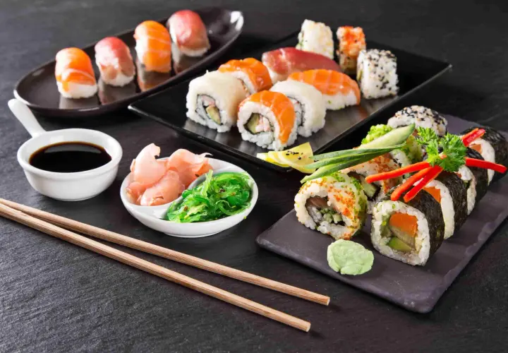 Różnorodne sushi, sos sojowy, marynowany imbir, sałatka wakame i pałeczki na ciemnym tle.