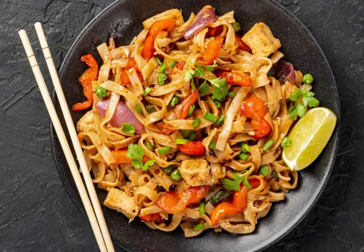 Pad thai z tofu, warzywami i limonką na czarnym talerzu, obok pałeczki.