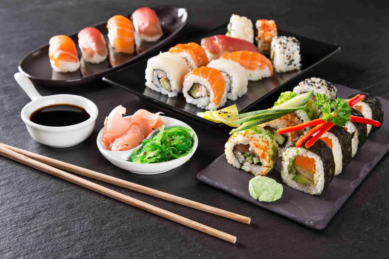 Różnorodne sushi, sos sojowy, marynowany imbir, sałatka wakame i pałeczki na ciemnym tle.