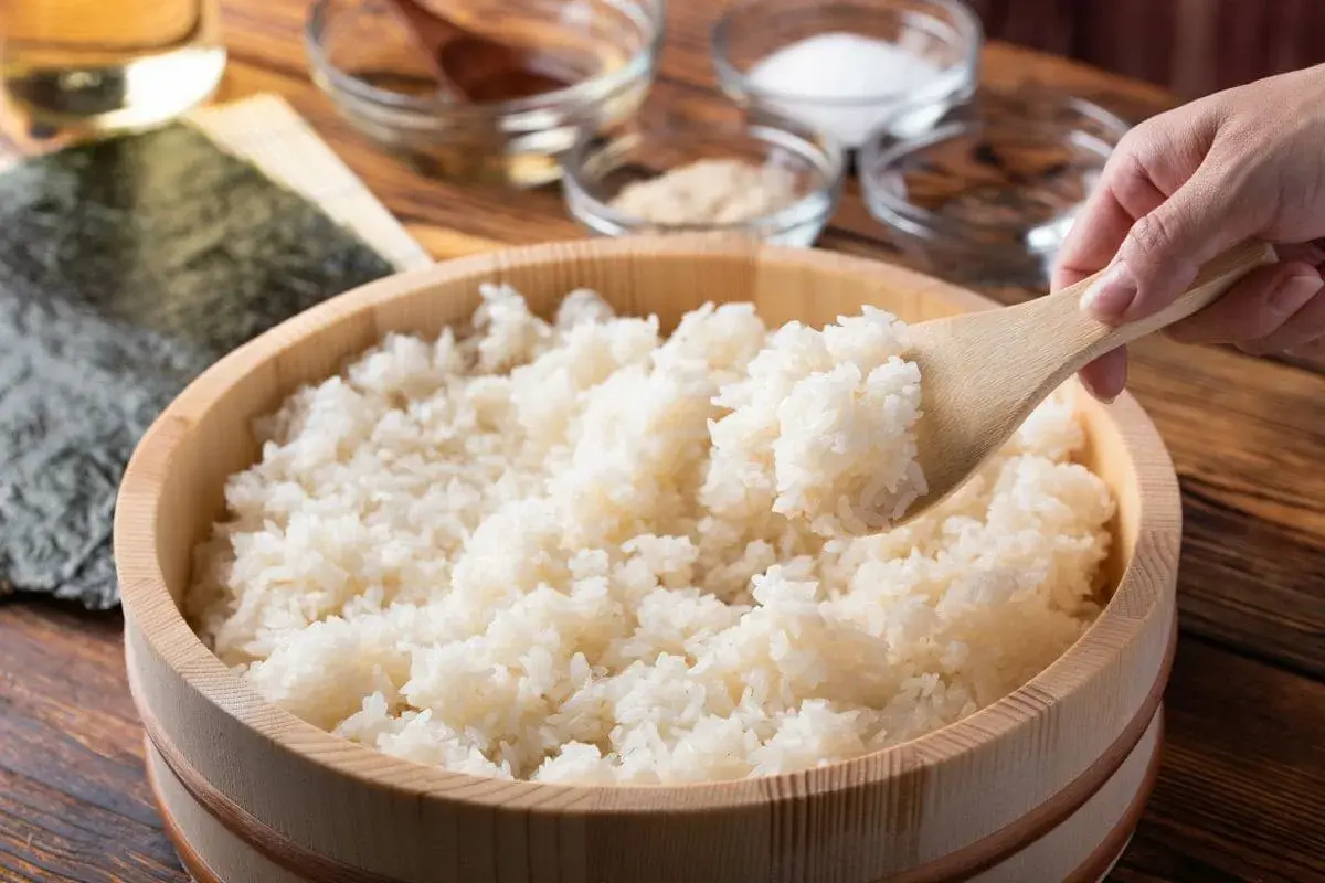 Drewniana miska z ryżem do sushi, nabierana drewnianą łyżką. W tle składniki.
