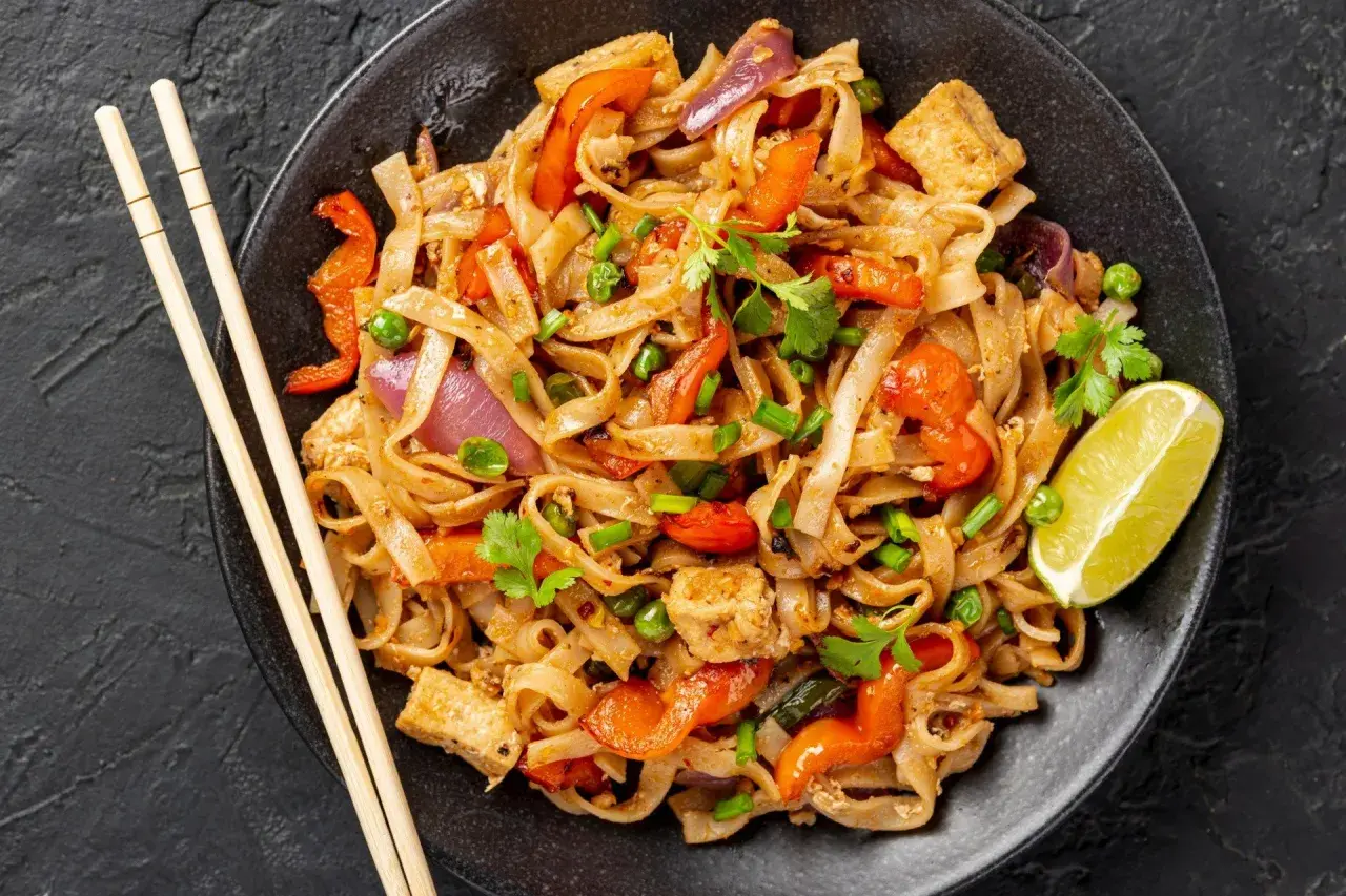 Pad thai z tofu, warzywami i limonką na czarnym talerzu, obok pałeczki.